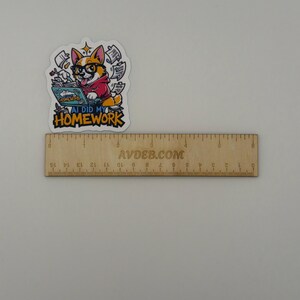 May include: A die-cut sticker featuring a cartoon corgi wearing glasses and a red hoodie, typing on a laptop with papers flying around. The text reads AI DID MY HOMEWORK. A wooden ruler is placed below the sticker, showing it is about 6.5 cm wide.