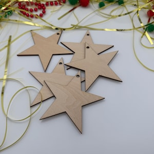 May include: A set of five flat, light-colored wooden star-shaped ornaments. Each star features a smooth, natural wood grain finish and a small circular hole at the top point, designed for hanging. The stars are arranged in a loose, overlapping cluster.