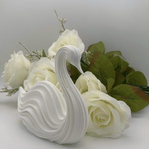 May include: A white swan figurine featuring a smooth, curved, and layered design that mimics the appearance of feathers. The swan is positioned in front of a cluster of white artificial roses with green leaves and small white filler flowers.