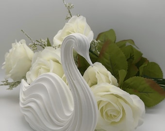 May include: A white swan figurine featuring a smooth, curved, and layered design that mimics the appearance of feathers. The swan is positioned in front of a cluster of white artificial roses with green leaves and small white filler flowers.