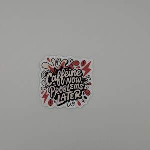 Puede incluir: Una pegatina troquelada con el texto negro Caffeine NOW, PROBLEMS LATER en un estilo de letra a mano audaz. El dise&ntilde;o est&aacute; acentuado con salpicaduras abstractas rojas y crema, formas de rayos y peque&ntilde;os iconos de granos de caf&eacute;.
