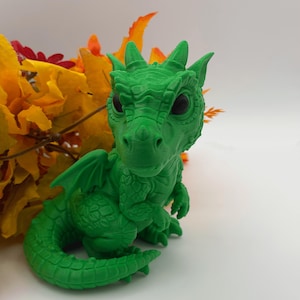 Puede incluir: Una figura de dragón de color verde brillante con grandes ojos oscuros, escamas texturizadas, alas pequeñas y cuernos en la cabeza. La criatura está representada en posición sentada con su larga cola curva envuelta alrededor de su cuerpo.
