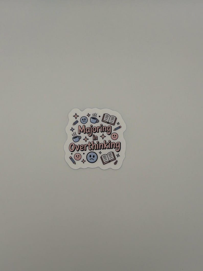 May include: A die-cut sticker featuring the text Majoring in Overthinking in a pink and brown font. The design includes blue coffee mugs, open books, pencils, four-pointed stars, and blue and pink sad face emojis scattered around the lettering.