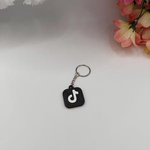 May include: A black square keyring featuring the white TikTok logo in the centre. The keyring is attached to a silver metal split ring by a short silver chain. The design is simple and features a raised white musical note icon on a black base.