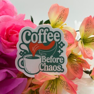 May include: A die-cut sticker featuring a white coffee mug with a dark green outline and a red liquid splash. The text reads Coffee Before Chaos in dark green retro-style lettering, accented with small star shapes against a white background.