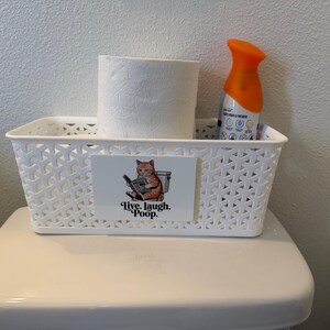 May include: A white rectangular plastic storage basket with a lattice pattern. A rectangular sign on the front features an illustration of an orange cat sitting on a toilet reading a newspaper with the text Live. Laugh. Poop. below the cat.