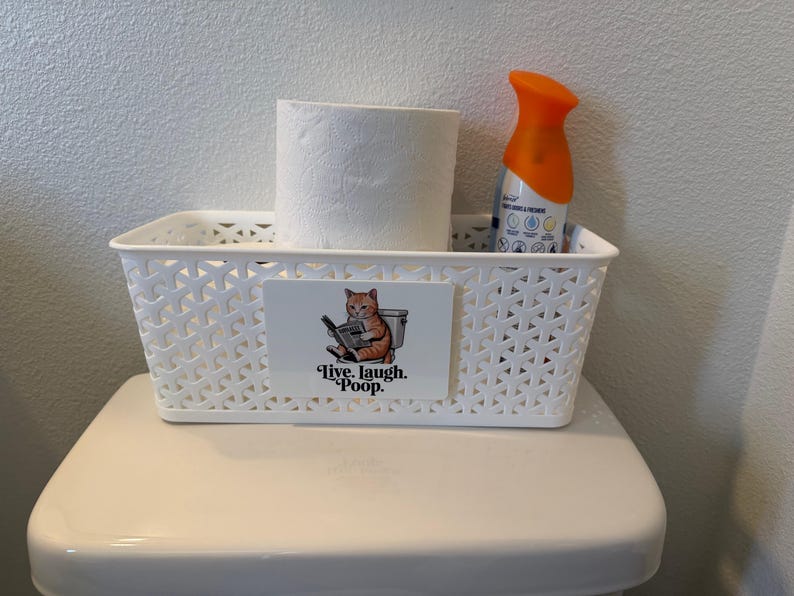 May include: A white rectangular plastic storage basket with a lattice pattern. A rectangular sign on the front features an illustration of an orange cat sitting on a toilet reading a newspaper with the text Live. Laugh. Poop. below the cat.