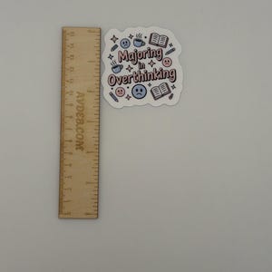 May include: A die-cut sticker with the text "Majoring in Overthinking" in pink and brown letters, surrounded by icons of books, coffee cups, pencils, and sad faces. A wooden ruler is placed next to it, showing the sticker is approximately 2.5 inches wide.