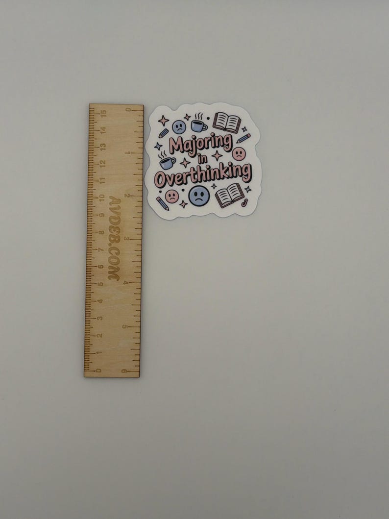 May include: A die-cut sticker with the text "Majoring in Overthinking" in pink and brown letters, surrounded by icons of books, coffee cups, pencils, and sad faces. A wooden ruler is placed next to it, showing the sticker is approximately 2.5 inches wide.
