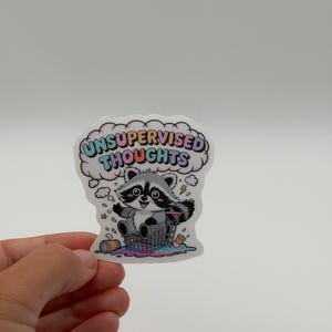 May include: A die-cut sticker featuring a cartoon raccoon sitting in a shopping cart with a pink, blue, and yellow thought bubble above its head containing the text UNSUPERVISED THOUGHTS in colorful letters, surrounded by scattered trash items.