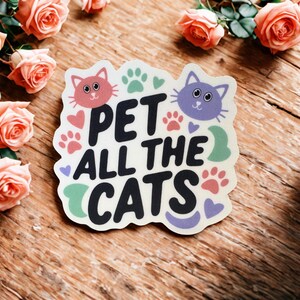 May include: A sticker with the text "PET ALL THE CATS" in black, with cat faces, paw prints, and hearts. The sticker is surrounded by peach-colored roses on a wooden surface.