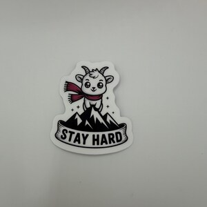 May include: A die-cut sticker featuring a white cartoon goat wearing a maroon scarf, standing atop a black and white mountain peak. Below the mountain, a banner contains the black text STAY HARD in a bold, sans-serif capital letter font.