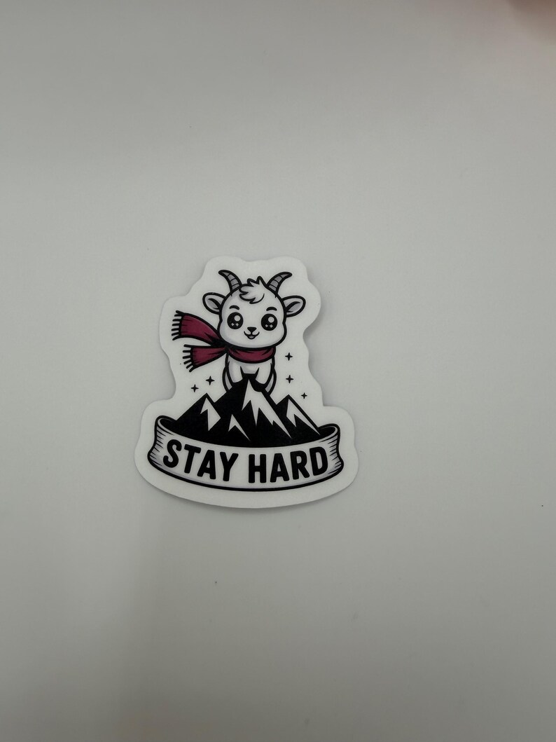 May include: A die-cut sticker featuring a white cartoon goat wearing a maroon scarf, standing atop a black and white mountain peak. Below the mountain, a banner contains the black text STAY HARD in a bold, sans-serif capital letter font.