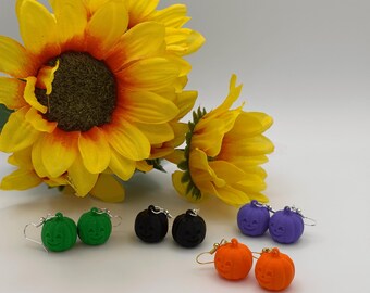 May include: Four pairs of pumpkin-shaped earrings in green, black, purple, and orange. Each pair features a carved jack-o'-lantern face and is attached to a metal earring hook. The earrings are arranged in a group with bright yellow sunflowers.