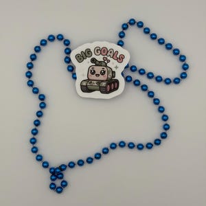 Puede incluir: Un collar hecho de cuentas esf&eacute;ricas de color azul brillante que presenta una pegatina blanca con un personaje de tanque de dibujos animados. El tanque tiene ojos grandes, una peque&ntilde;a sonrisa y dos corazones rosas encima. El texto BIG GOALS est&aacute; impreso.