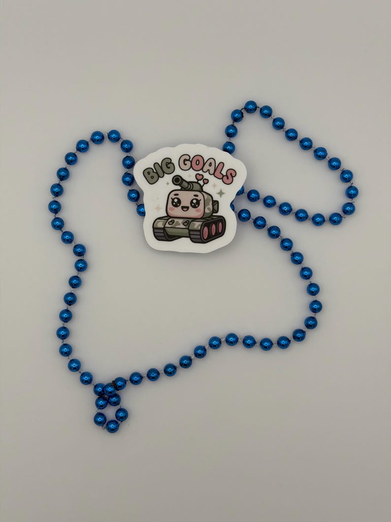 Puede incluir: Un collar hecho de cuentas esf&eacute;ricas de color azul brillante que presenta una pegatina blanca con un personaje de tanque de dibujos animados. El tanque tiene ojos grandes, una peque&ntilde;a sonrisa y dos corazones rosas encima. El texto BIG GOALS est&aacute; impreso.