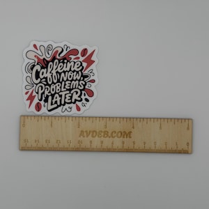 Puede incluir: Una pegatina troquelada con el texto negro "CAFFEINE NOW, PROBLEMS LATER" rodeado de formas abstractas rojas y negras. Una regla de madera se sit&uacute;a debajo de la pegatina, mostrando una anchura de aproximadamente 6,5 cm.