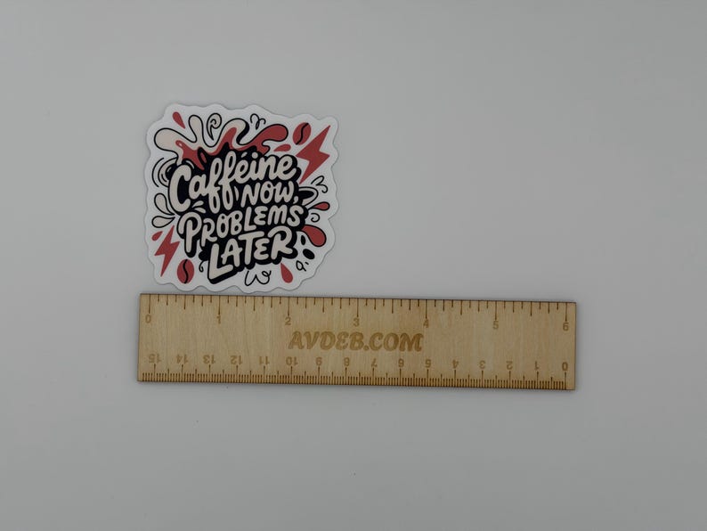 Puede incluir: Una pegatina troquelada con el texto negro "CAFFEINE NOW, PROBLEMS LATER" rodeado de formas abstractas rojas y negras. Una regla de madera se sit&uacute;a debajo de la pegatina, mostrando una anchura de aproximadamente 6,5 cm.