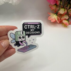 May include: A die-cut sticker featuring a light green floppy disk character with wings, typing on a keyboard. A speech bubble above the character contains the black text CTRL+Z MY DECISIONS. The design includes small sparkles and a purple gradient.