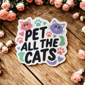 May include: A sticker with the text "PET ALL THE CATS" in black, with a pink cat face and paw prints. The sticker is surrounded by pink roses on a wooden surface.