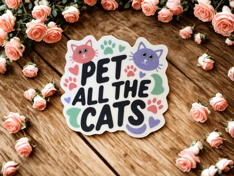 May include: A sticker with the text "PET ALL THE CATS" in black, with a pink cat face and paw prints. The sticker is surrounded by pink roses on a wooden surface.