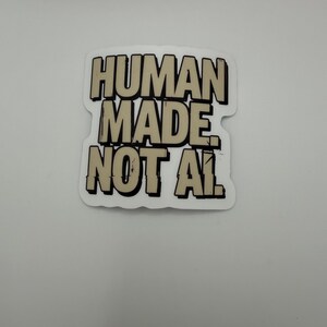 May include: A die-cut sticker featuring the text HUMAN MADE NOT AI in bold, blocky, beige-colored letters with thick black outlines and drop shadows. The design has a slightly distressed, comic-book style aesthetic with a white border.
