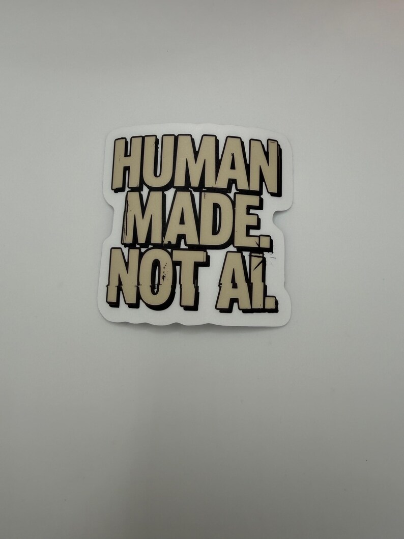 May include: A die-cut sticker featuring the text HUMAN MADE NOT AI in bold, blocky, beige-colored letters with thick black outlines and drop shadows. The design has a slightly distressed, comic-book style aesthetic with a white border.