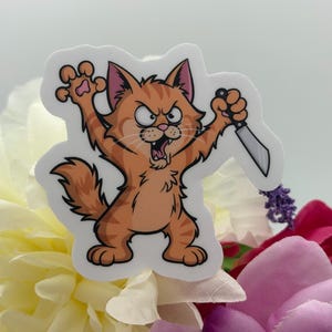 May include: A die-cut vinyl sticker featuring a cartoon orange tabby cat with a fierce expression, baring its teeth and claws. The cat is standing on its hind legs, holding a small silver kitchen knife in one paw and raising the other paw.