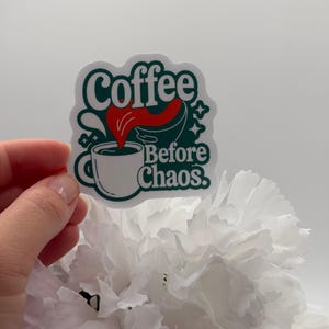 May include: A die-cut sticker featuring a white coffee mug with dark green outlines and a red liquid splash. The text reads Coffee Before Chaos in dark green lettering. The design includes small star accents and a dark green background shape.