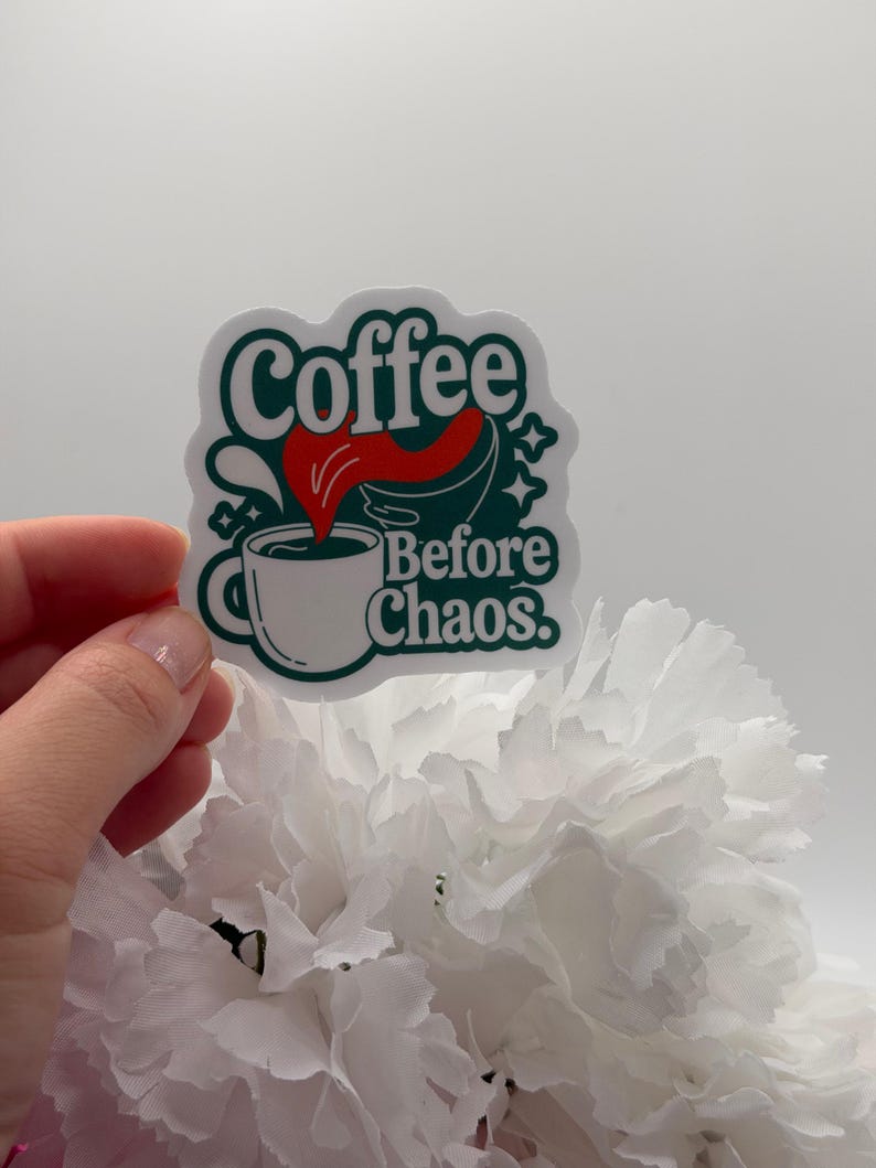 May include: A die-cut sticker featuring a white coffee mug with dark green outlines and a red liquid splash. The text reads Coffee Before Chaos in dark green lettering. The design includes small star accents and a dark green background shape.