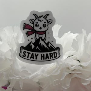 May include: A die-cut sticker featuring a cartoon goat with a maroon scarf standing atop a black mountain peak. The design includes small star accents and a banner at the bottom with the text STAY HARD in bold black capital letters.