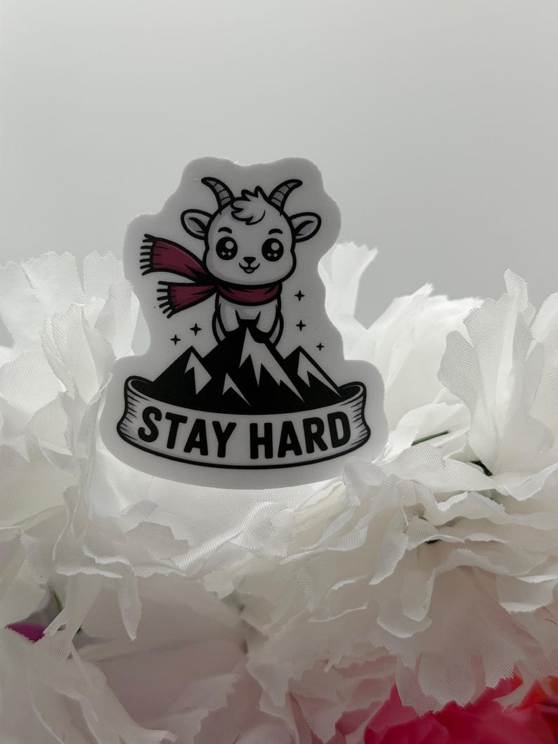 May include: A die-cut sticker featuring a cartoon goat with a maroon scarf standing atop a black mountain peak. The design includes small star accents and a banner at the bottom with the text STAY HARD in bold black capital letters.