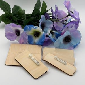 May include: A collection of rectangular light-colored wooden name badge blanks with white plastic pin backings attached to the reverse side, arranged in a cluster beneath a bouquet of artificial purple and blue flowers with green leaves.