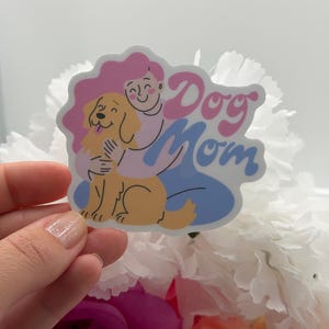 Puede incluir: Una pegatina troquelada con una ilustraci&oacute;n de dibujos animados de una persona con pelo rosa abrazando a un perro de color dorado. El texto "Dog Mom" est&aacute; escrito en una fuente rosa, negrita y redondeada. La persona viste una camisa morada clara.