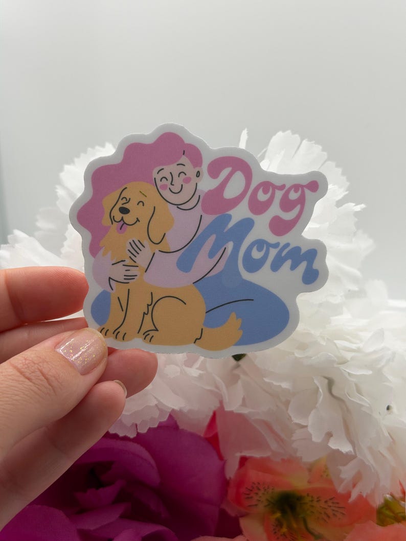 Puede incluir: Una pegatina troquelada con una ilustraci&oacute;n de dibujos animados de una persona con pelo rosa abrazando a un perro de color dorado. El texto "Dog Mom" est&aacute; escrito en una fuente rosa, negrita y redondeada. La persona viste una camisa morada clara.