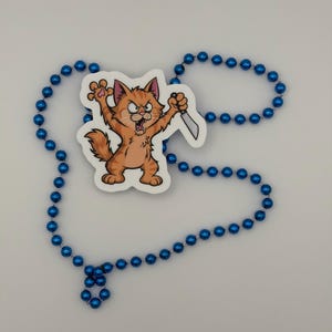 May include: A sticker featuring an orange tabby cat with an angry expression, standing on its hind legs with claws extended and holding a silver knife, surrounded by a long strand of shiny blue plastic beads arranged in a loose circular shape.
