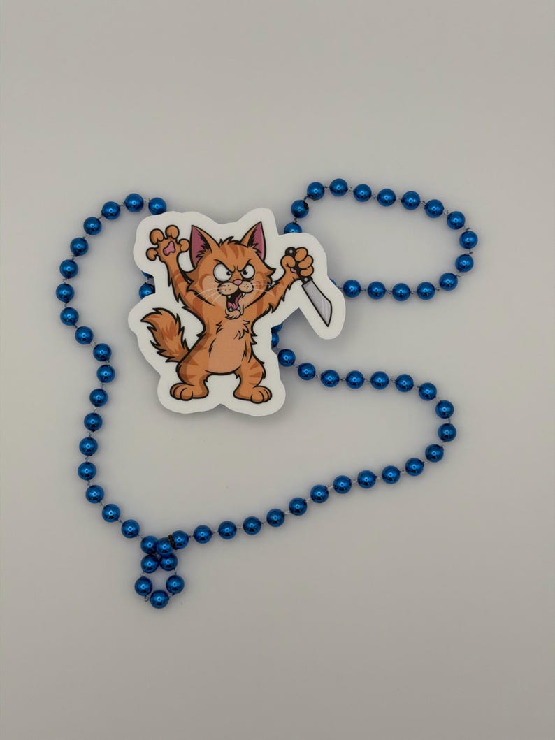 May include: A sticker featuring an orange tabby cat with an angry expression, standing on its hind legs with claws extended and holding a silver knife, surrounded by a long strand of shiny blue plastic beads arranged in a loose circular shape.
