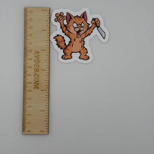 May include: An orange tabby cat sticker with a cartoon design, featuring an angry expression, raised claws, and holding a silver knife. The sticker is positioned next to a wooden ruler showing measurements in both inches and centimetres.