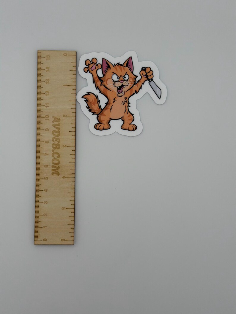 May include: An orange tabby cat sticker with a cartoon design, featuring an angry expression, raised claws, and holding a silver knife. The sticker is positioned next to a wooden ruler showing measurements in both inches and centimetres.