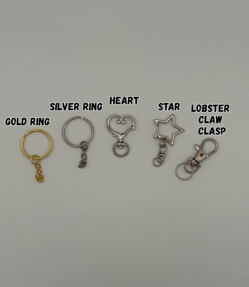 May include: A collection of metal keychain hardware including a gold ring, a silver ring, a heart-shaped clasp, a star-shaped clasp, and a lobster claw clasp. Each item is labeled with its corresponding name in black text above the hardware.
