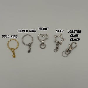 May include: A collection of metal keychain hardware including a gold ring, a silver ring, a heart-shaped clasp, a star-shaped clasp, and a lobster claw clasp. Each item is labeled with its corresponding name in black text above the hardware.