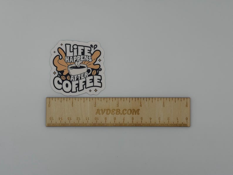 May include: A die-cut sticker featuring a coffee cup graphic with the text "LIFE HAPPENS AFTER COFFEE" in black and tan colours. The sticker is positioned above a wooden ruler that shows a measurement of approximately 7.5 cm in width.