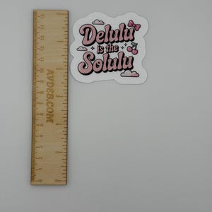 May include: A die-cut sticker featuring the pink and black text "Delulu is the Solulu" with small pink cherry and cloud illustrations. A wooden ruler is placed next to the sticker, showing it measures approximately 6.5 cm in width.