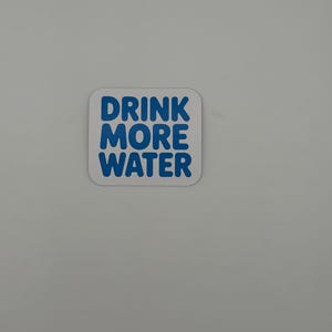 May include: A square sticker with rounded corners featuring a white background and the text DRINK MORE WATER printed in bold, blue, sans-serif capital letters. The sticker has a clean, simple design with the text centered on the surface.