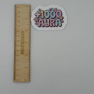 May include: A die-cut sticker featuring the text +1000 AURA in a bubbly, pastel-colored font with small star and heart accents, positioned next to a wooden ruler showing a width of approximately 3 inches and a height of 2 inches.