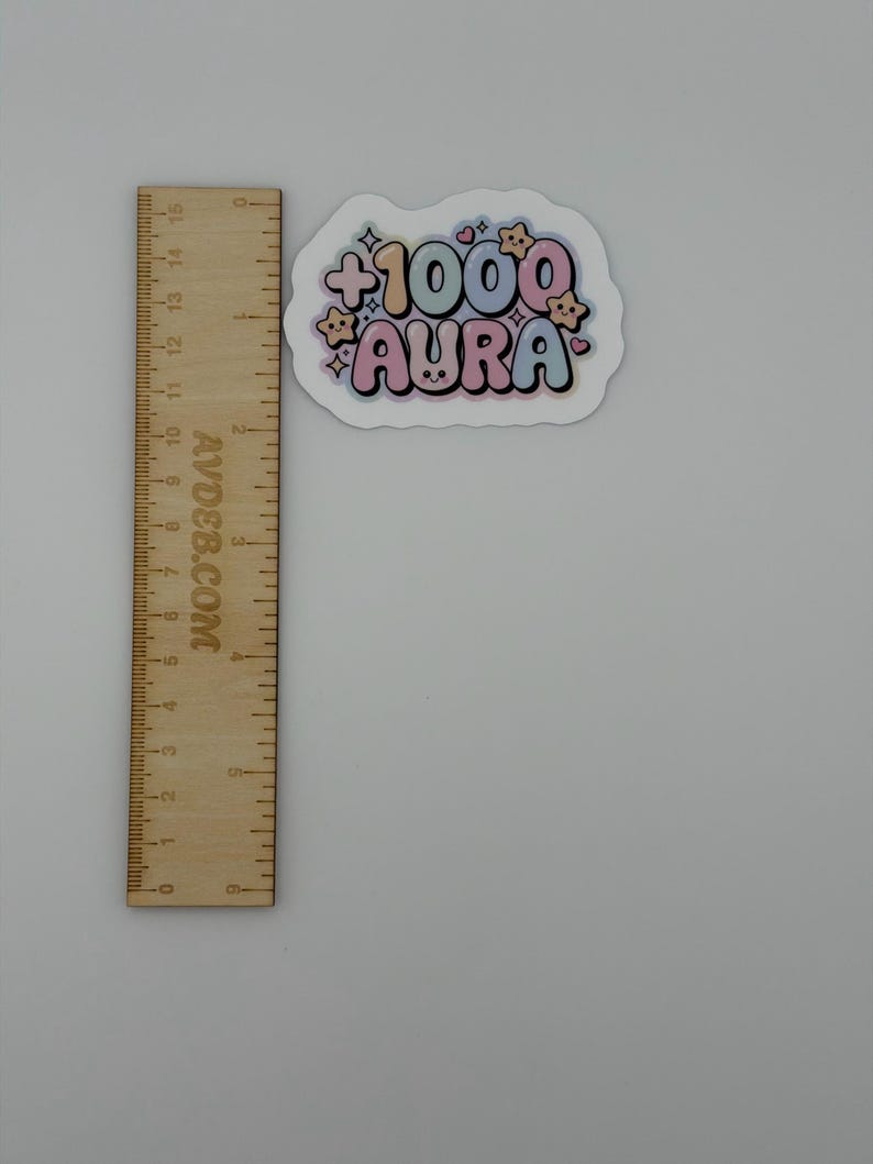 May include: A die-cut sticker featuring the text +1000 AURA in a bubbly, pastel-colored font with small star and heart accents, positioned next to a wooden ruler showing a width of approximately 3 inches and a height of 2 inches.