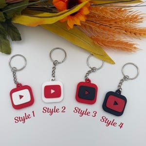 May include: Four square keychains featuring a play button icon, labeled Style 1, Style 2, Style 3, and Style 4. The keychains come in color combinations of red and white or red and black, each attached to a silver metal split ring by a short chain.