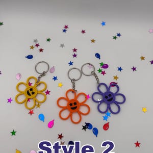 Peut inclure: Trois porte-cl&eacute;s en forme de fleur de couleur jaune, orange et violette, chacun dot&eacute; d'un visage souriant central avec des yeux en forme d'&eacute;toile et un anneau m&eacute;tallique. Le texte "Style 2" est imprim&eacute; en grandes lettres violettes en bas.