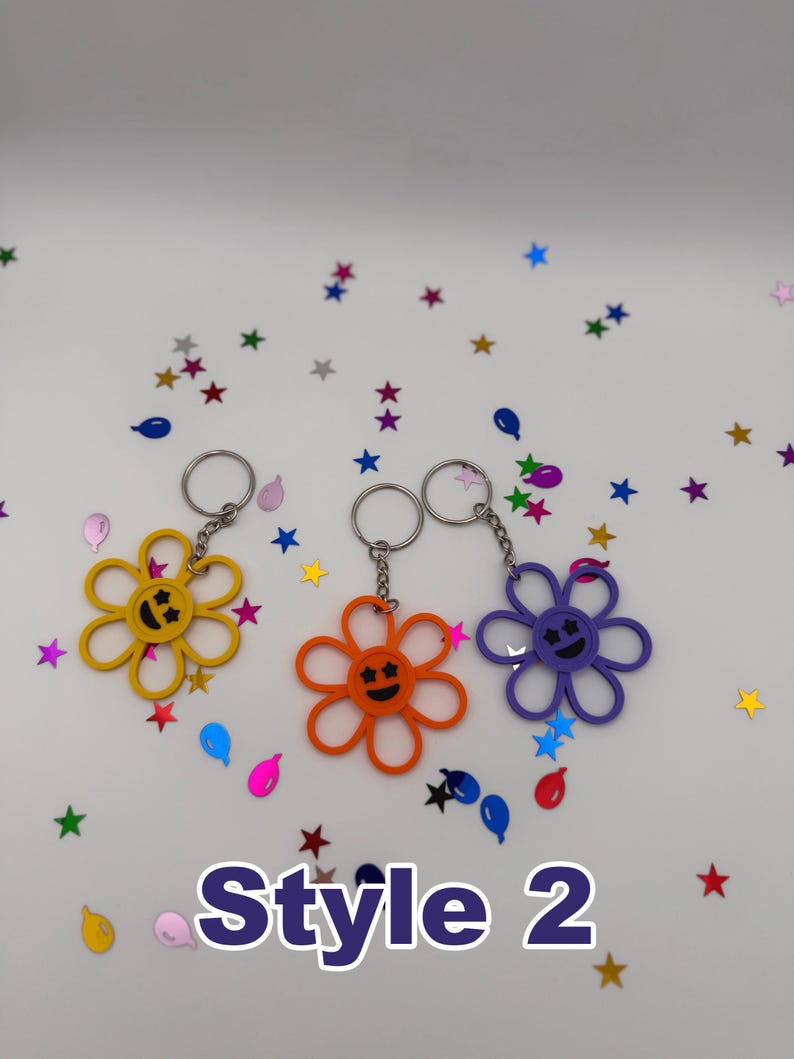 Peut inclure: Trois porte-cl&eacute;s en forme de fleur de couleur jaune, orange et violette, chacun dot&eacute; d'un visage souriant central avec des yeux en forme d'&eacute;toile et un anneau m&eacute;tallique. Le texte "Style 2" est imprim&eacute; en grandes lettres violettes en bas.
