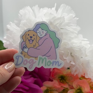 Op de afbeelding: Een gestanste sticker met een kleurrijke illustratie van een persoon met lichtblauw en groen haar in een roze trui, die een goudkleurige hond knuffelt. De tekst "Dog Mom" is onderaan de sticker gedrukt in een lichtpaars cursief lettertype.